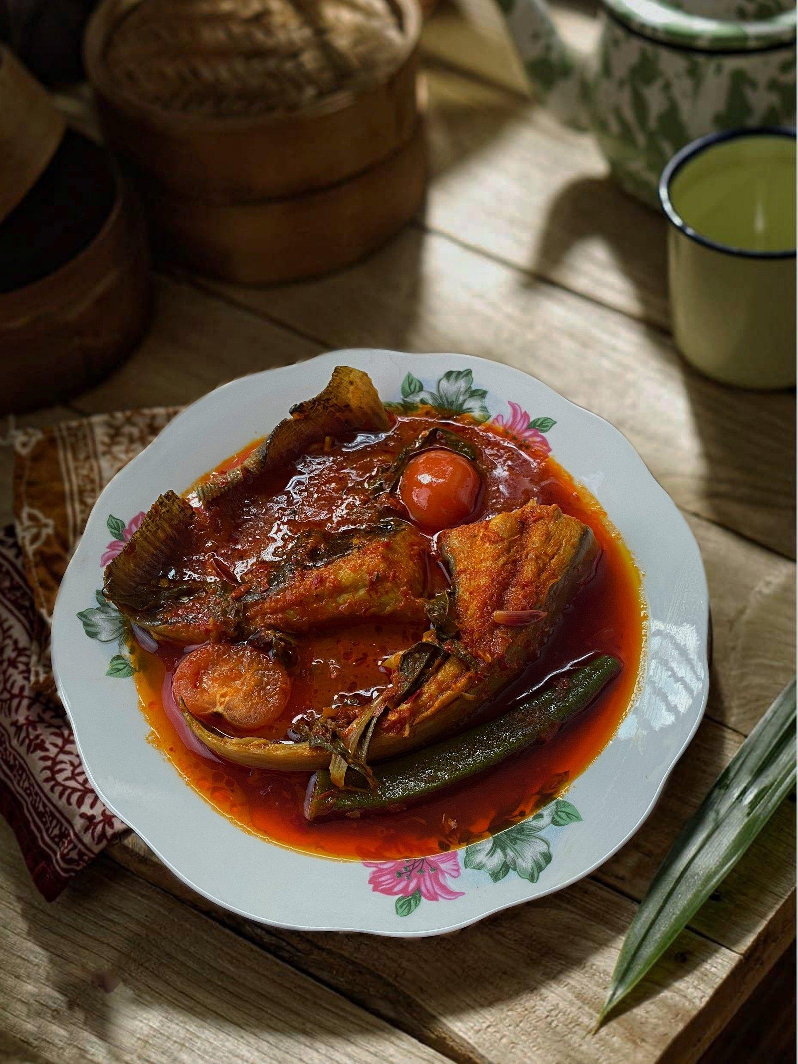 Asam pedas ikan pari — stingray wings in a scarlet tamarind gravy with okra, tomato and daun kesum, on a floral enamel plate.