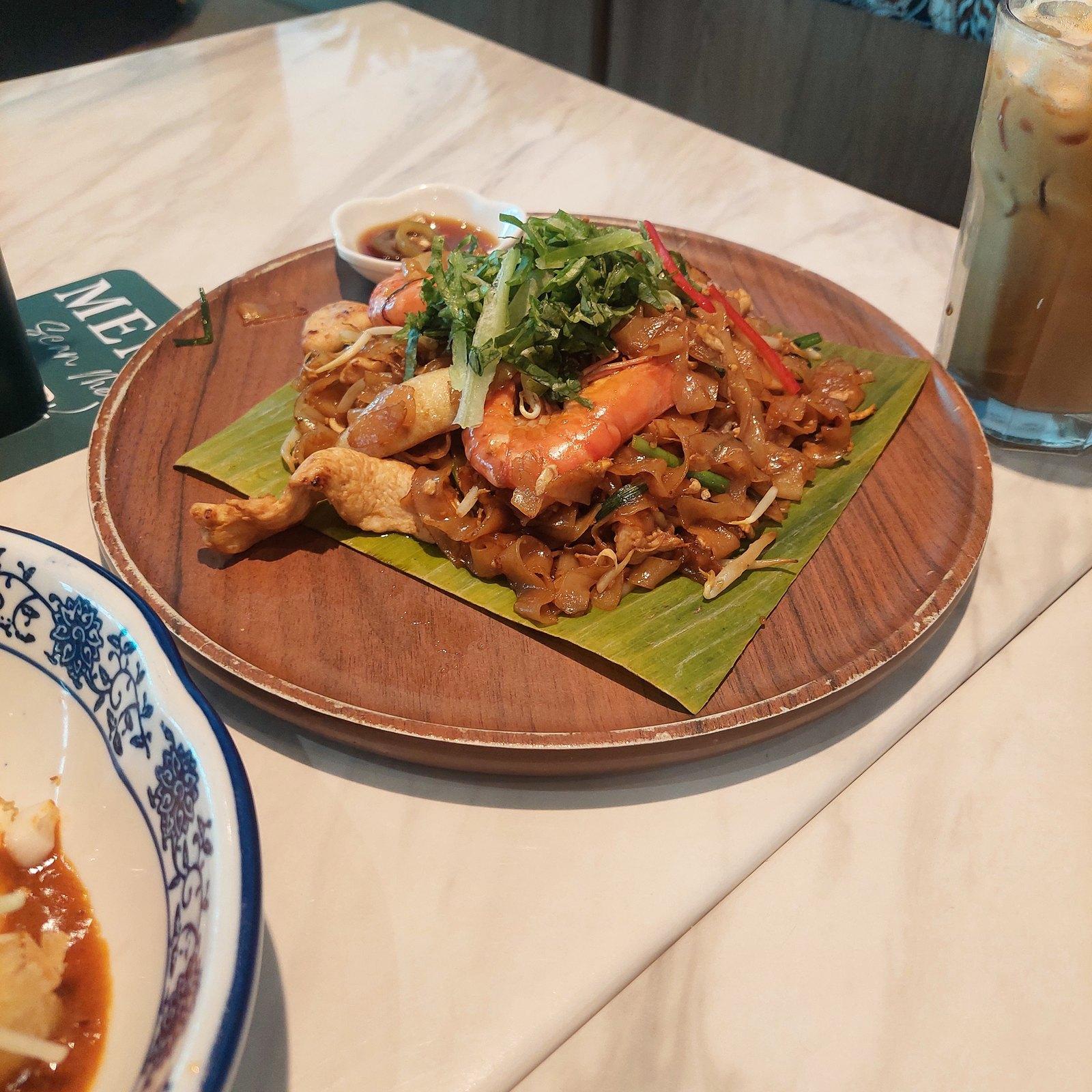 Char kway teow on a banana leaf — dark soy-stained flat rice noodles tossed with a whole prawn, bean sprouts and chives, served on a round wooden board.