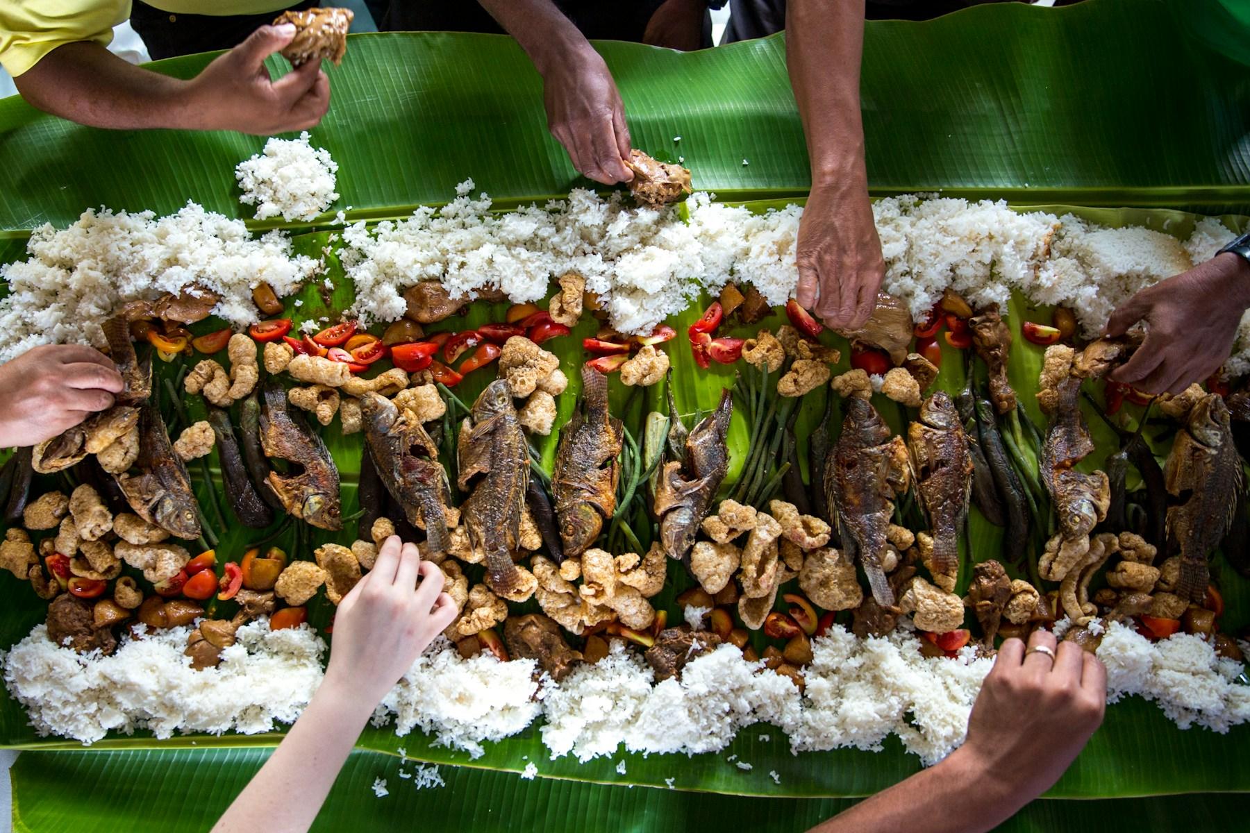 Many hands eating with their fingers from a long banana leaf spread of rice and side dishes.