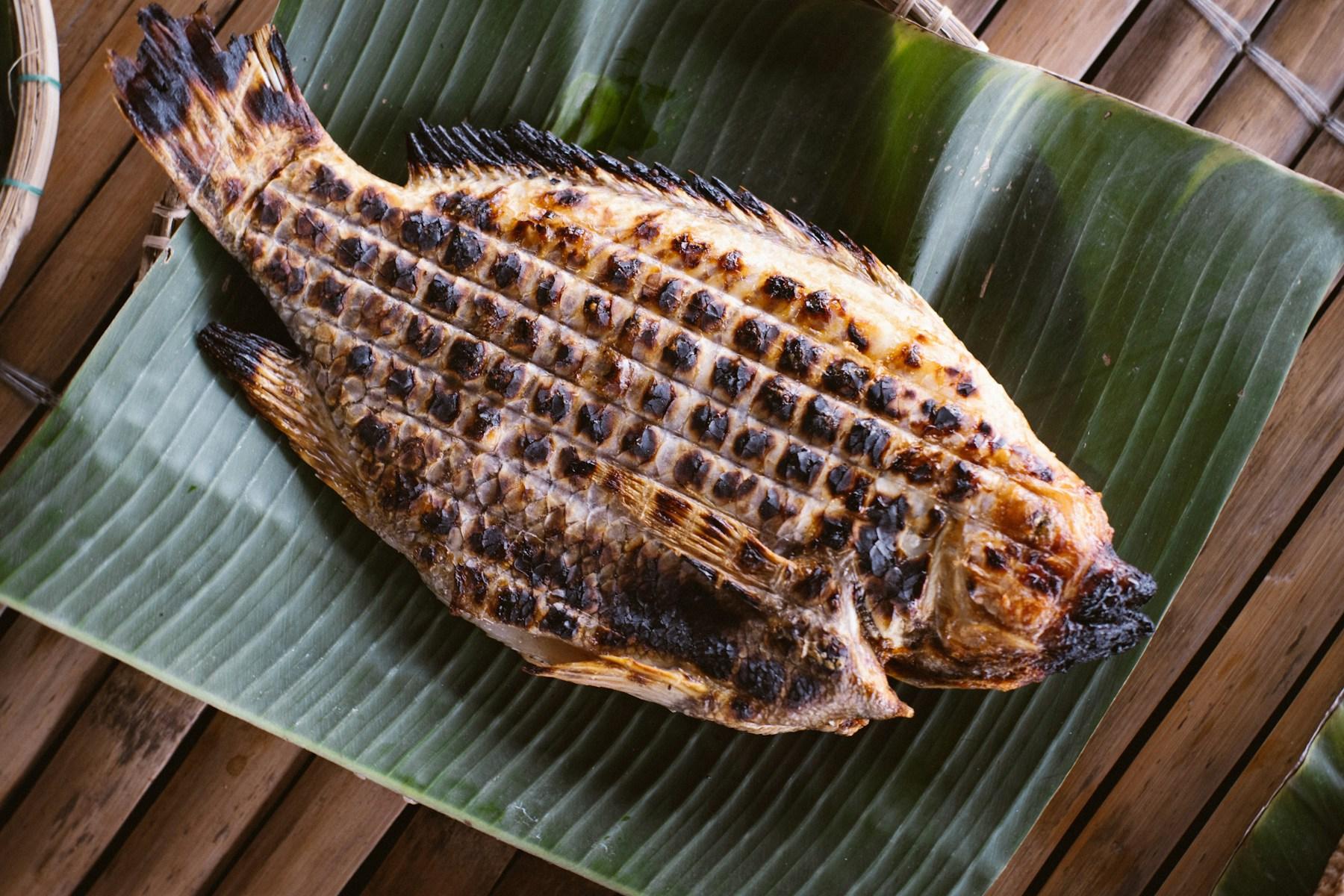 A whole fish grilled until charred and scored across the skin, resting on a banana leaf.