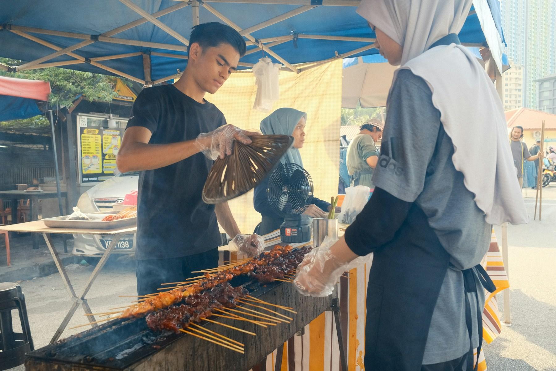A young cook fanning the coals with a woven fan over a long charcoal grill packed with satay skewers, a colleague turning them at the far end of an open-air stall.
