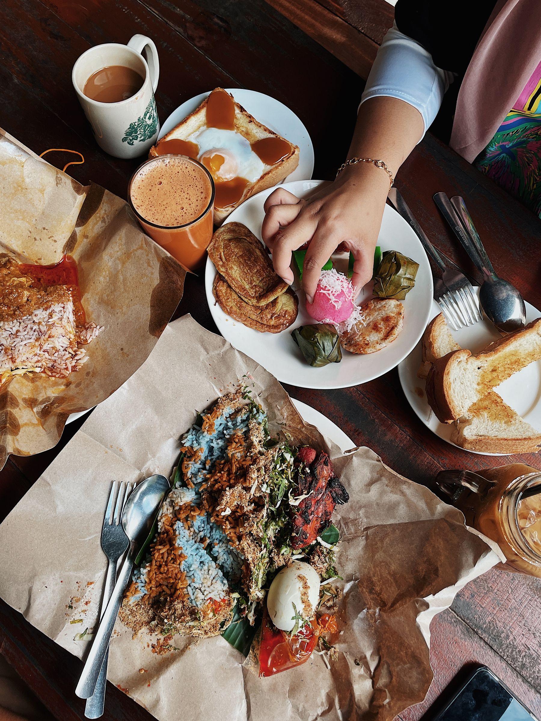 A shared Malaysian breakfast table seen from above: nasi kerabu on paper, nasi lemak in its wrapper, a plate of kuih and roti, kaya toast and glasses of teh tarik, with a hand reaching in.