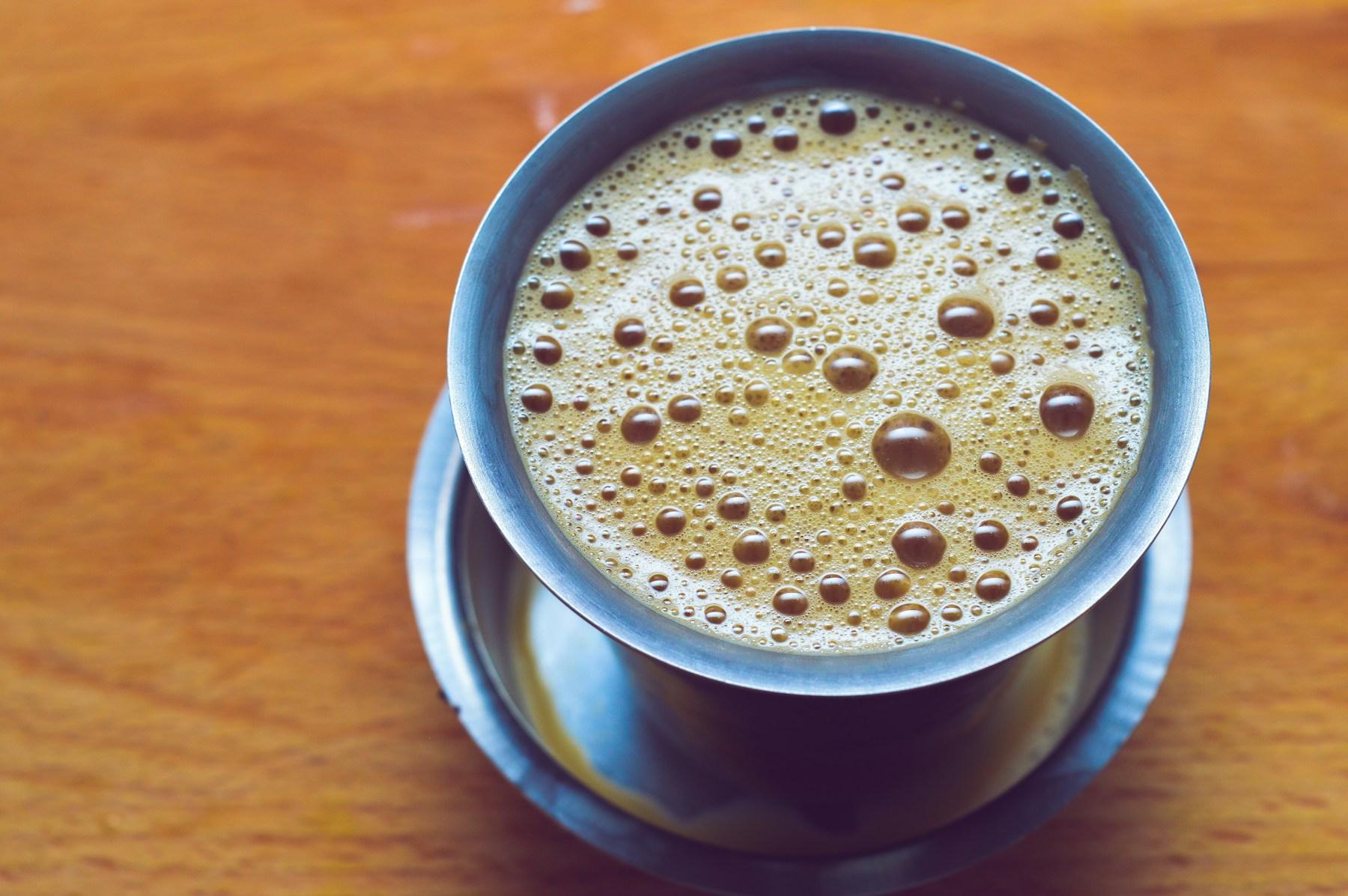 A stainless steel cup of milky pulled tea on its saucer, seen from above, the surface still bubbled from the pour.