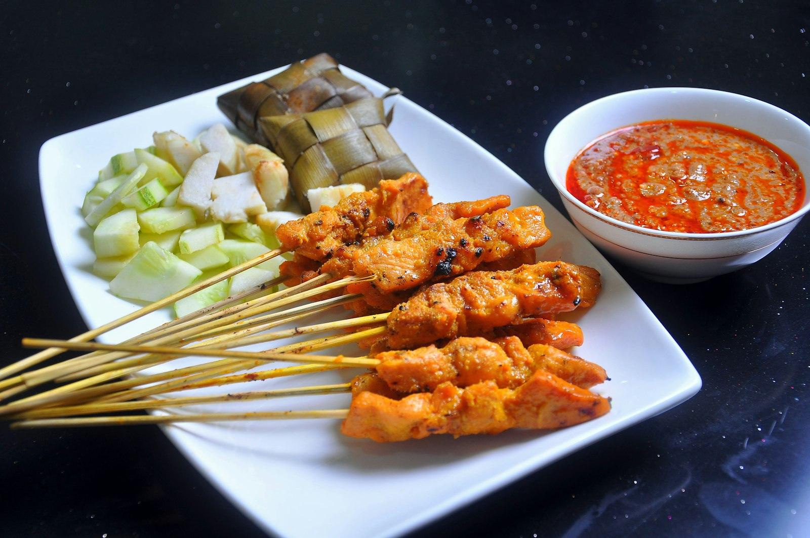 Grilled chicken satay skewers on a white plate with a bowl of peanut sauce, cucumber chunks and pressed rice cakes.