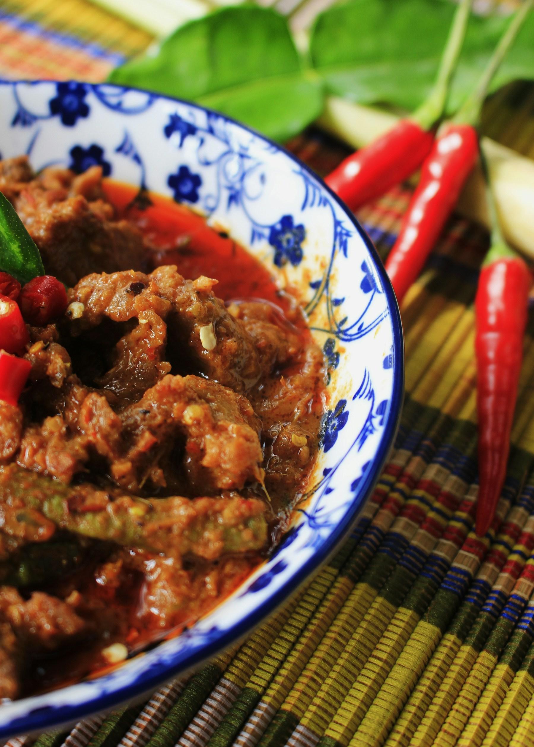 Dark, richly spiced beef rendang piled in a blue and white patterned bowl, garnished with red chillies and herbs.