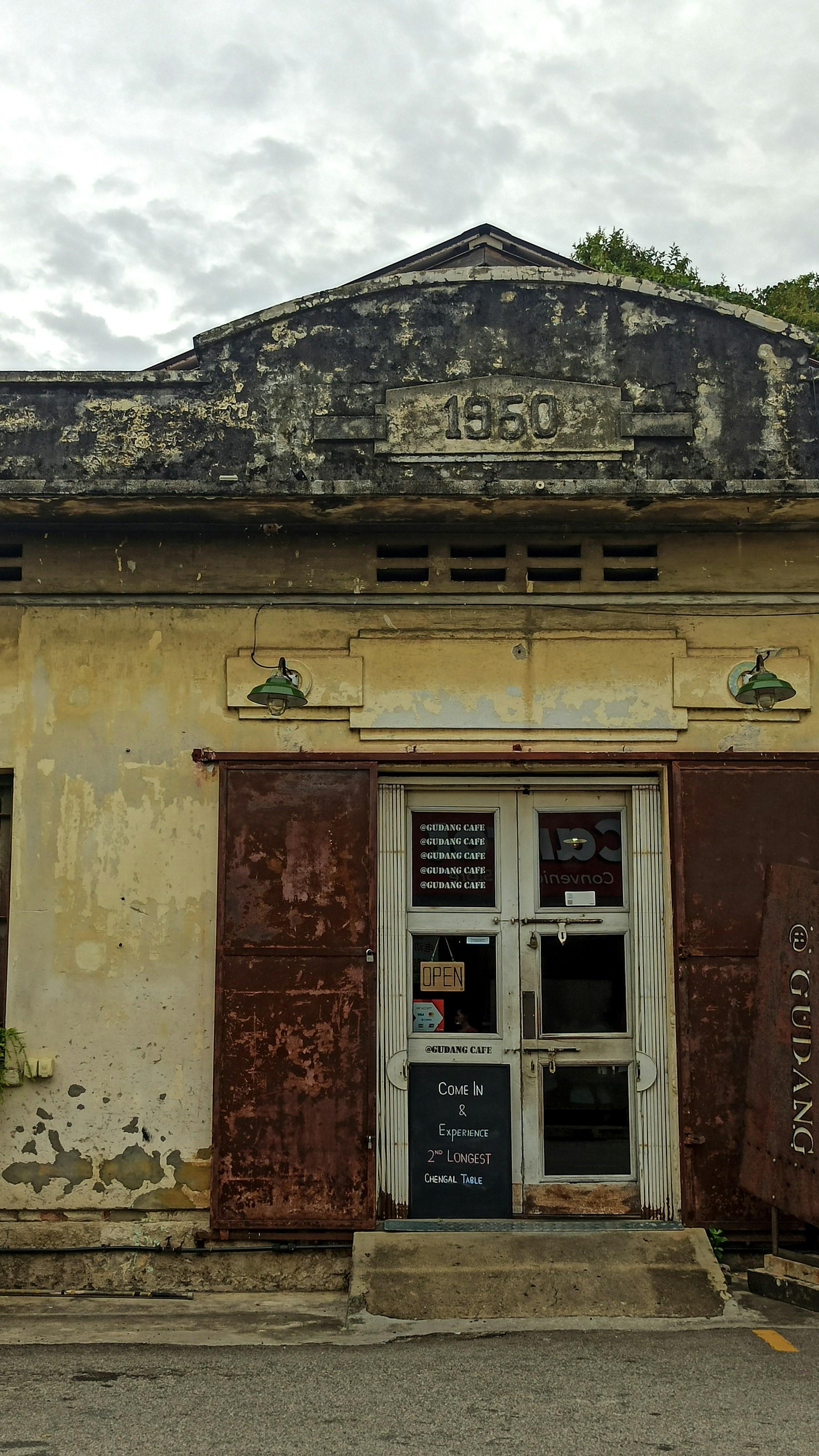 The weathered facade of a 1950 shophouse, pale plaster peeling around rusted steel shutters and a glass-panelled door.