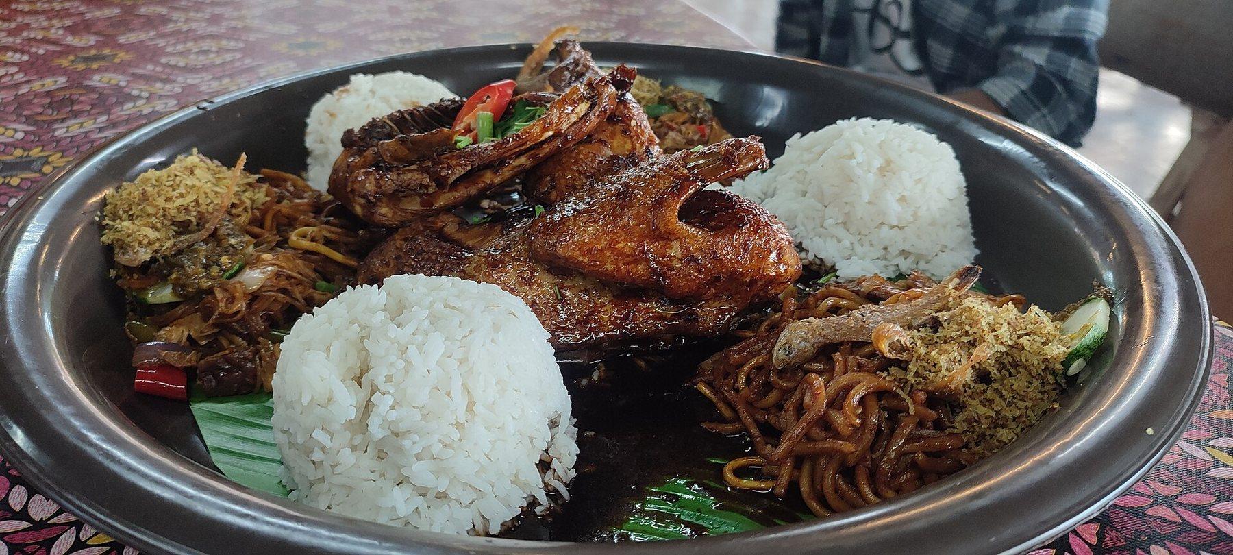 A wide steel tray of nasi ambeng lined with banana leaf - mounds of white rice, glazed fried chicken, mee goreng, salted fish, serunding and sambal, laid out to be shared.