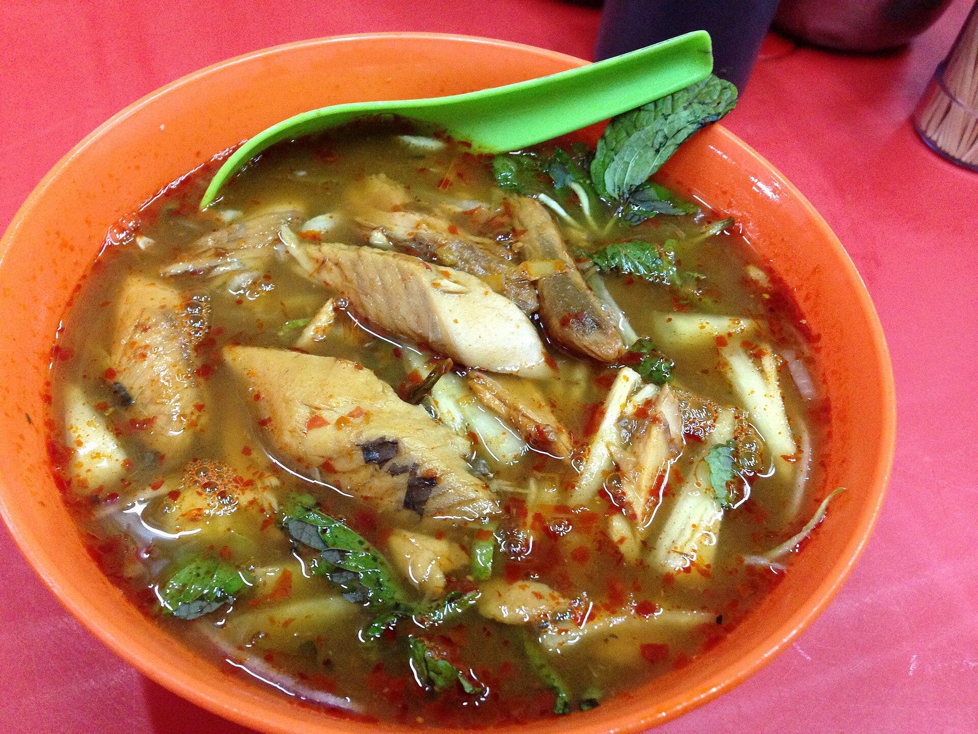A bowl of asam laksa: amber tamarind fish broth flecked with chilli, flaked mackerel, shredded cucumber and pineapple and sprigs of mint over thick white rice noodles.