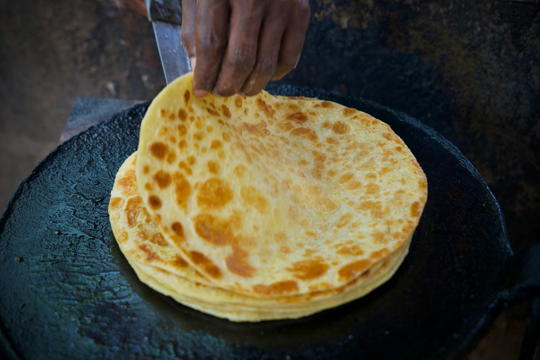 A hand lifting the top of a stack of golden, blistered flatbreads from a hot, oiled cast-iron griddle.